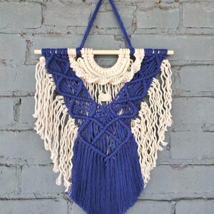 May include: A macramé wall hanging with a geometric design in blue and cream. The wall decor is suspended from a wooden dowel and has a triangular shape with fringe detailing. The background is a grey brick wall.