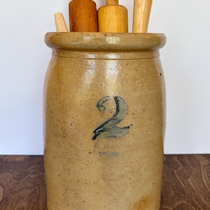 May include: A tan stoneware crock with a blue number 2 stamped on the front, containing four wooden kitchen tools of varying heights and shades. The cylindrical crock features a thick, rounded rim and a speckled, glazed finish throughout.