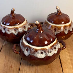 May include: Three round ceramic lidded bowls with two side handles each. The bowls feature a glossy dark brown glaze with a decorative white drip pattern around the upper rim. Each lid is shaped like a pumpkin with a small brown stem handle.