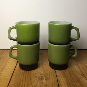 May include: Four stackable mugs made of green glass with a dark brown gradient at the base. Each mug features a cylindrical body, a smooth rim, and a square-shaped handle. The mugs are arranged in two vertical stacks of two, showcasing the color transition from light olive green at the top to a deep, solid brown at the bottom.