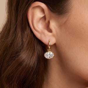 May include: A gold hoop earring featuring a small, oval-shaped clear gemstone charm suspended from the bottom. The earring is crafted from a polished gold-tone metal, with the faceted stone catching the light to create a bright, reflective appearance.