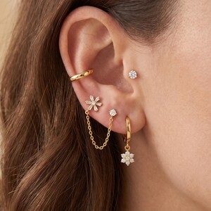 Könnte beinhalten: Ein goldener Ohrring-Cuff, ein kleiner runder Stecker und ein blumenförmiger Stecker, der durch eine feine goldene Kette mit einem zweiten Stecker verbunden ist. Ein goldener Creolen-Ohrring mit einem hängenden Blumenanhänger aus klaren Steinen.