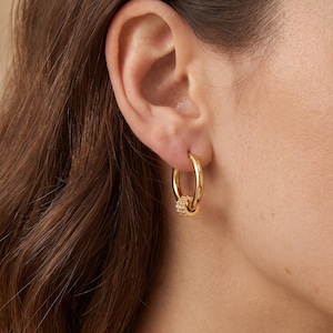 Könnte beinhalten: Ein goldener Creolen-Ohrring mit einem glatten, polierten kreisförmigen Band und einem kleinen, strukturierten Anhänger an der Unterseite. Der Anhänger hat eine zylindrische Form mit einer detaillierten, körnigen Oberfläche.