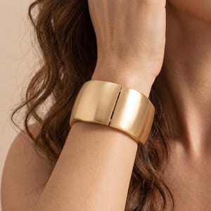 May include: A wide, matte gold-toned cuff bracelet with a smooth, curved surface and a vertical hinge closure mechanism. The bracelet features a solid, continuous band design that wraps around the wrist, showcasing a clean, metallic finish.