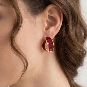 May include: A hoop earring featuring a deep red, glossy, semi-circular component nested within a thin, polished gold-tone metal frame. The design creates a layered, dimensional look with a smooth, reflective surface on the red portion.
