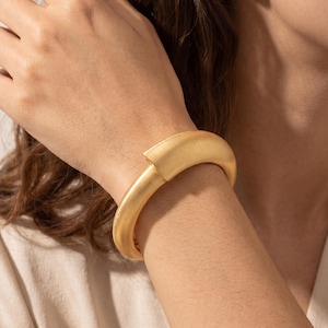 Può includere: Un bracciale rigido incernierato in color oro opaco con un design tubolare liscio e arrotondato. Il bracciale presenta un meccanismo di chiusura senza giunture e un aspetto solido e consistente che avvolge il polso con un profilo curvo.