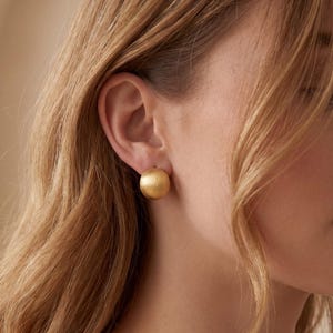 Può includere: Primo piano di un orecchino a bottone sferico color oro. L'orecchino ha una finitura opaca e viene indossato sull'orecchio di una persona con i capelli biondi. Lo sfondo è di un tono morbido e neutro.