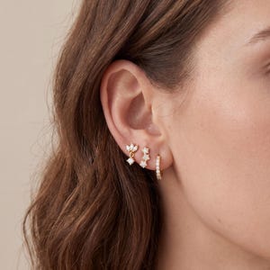 Könnte beinhalten: Drei goldfarbene Ohrringe an einem Ohr. Der erste ist ein kleiner Hänger mit einem marquise-geschliffenen Stein, der zweite ein linearer Stecker mit drei kleinen runden Steinen und der dritte ein kleiner Creolen-Ohrring mit kleinen klaren Steinen.
