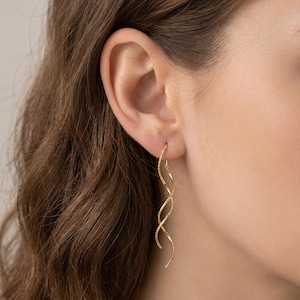 May include: A close-up of a person's ear wearing a pair of gold spiral earrings. The earrings have a textured surface and a curved design, creating a flowing, elegant look. The earrings are hanging from the ear.