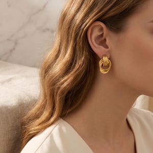 Peut inclure: Boucles d'oreilles dorées avec un motif tissé. Les boucles d'oreilles sont circulaires et comportent plusieurs bandes entrelacées, créant un aspect texturé et élégant. Les boucles d'oreilles sont de couleur or vif et se portent à l'oreille.