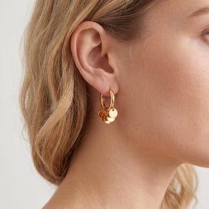 Puede incluir: Pendientes de aro dorados con colgantes circulares. Los pendientes son de un oro brillante y se llevan en la oreja de una persona. Los colgantes son pequeños discos planos que cuelgan de la parte inferior de los aros.