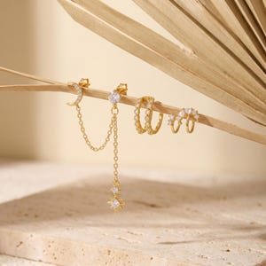 Puede incluir: Un conjunto de pendientes dorados que presenta un dije de media luna y estrella conectados por cadenas finas, junto a dos aros pequeños adornados con cristales transparentes y una perla blanca, todo con un acabado metálico pulido.