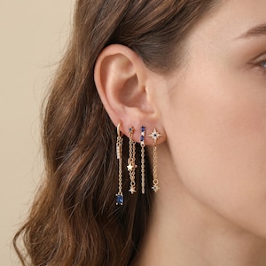 May include: A set of four gold-toned earrings featuring dangling chains. The designs include a baguette-cut blue stone, a star-shaped charm, a vertical bar with blue stones, and a starburst motif with a central dark stone, all worn in a single ear.
