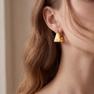 May include: Gold-coloured, triangular hoop earrings. The earrings have a flat, polished surface and feature the engraved text "FOREVER LOVE". The earrings are worn on the ear and are a simple, elegant design.