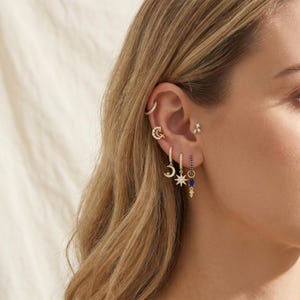 Könnte beinhalten: Eine Kollektion goldfarbener Ohrringe mit einem kleinen Creolen-Ohrring, einem Mondsichel-Anhänger, einem Stern-Anhänger und einem Hängeohrring mit einem blauen tropfenförmigen Stein. Das Set enthält eine kleine Ohrklemme und einen Ohrstecker mit drei Steinen.