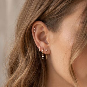 Könnte beinhalten: Ein Set aus goldenen Ohrringen mit einem kleinen Perlenstecker am oberen Ohr, einem dünnen Creolen-Ring mit Pavé-Besatz, einem goldenen Ring mit hängender Perle und kleinem Kristall sowie einem Stecker mit feiner Goldkette am Ohrläppchen.
