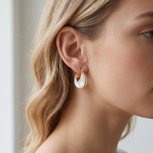 May include: Gold-tone hoop earrings with a crescent moon shape. The earrings feature a white, iridescent inlay and a polished gold finish. The earrings are shown on a person's ear.