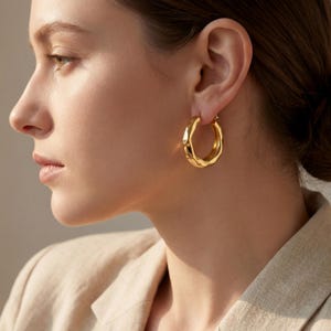 Puede incluir: Pendientes de aro dorados con un diseño texturizado y orgánico. Los pendientes son de tamaño mediano y tienen un acabado brillante. Los aros se llevan en la oreja de una persona que lleva una blazer beige.