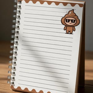 May include: A spiral-bound notebook with lined pages and a brown cover. A cartoon illustration of a smiling poop emoji wearing sunglasses is affixed to the top right corner. The notebook is open and standing upright on a wooden surface.