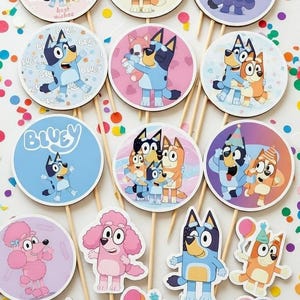 Puede incluir: Una colección de adornos para pasteles con el tema de Bluey sobre palitos de madera. Los adornos presentan varios personajes y frases como "Happy Birthday" y "Make a Wish" en un estilo de dibujos animados colorido. Hay confeti esparcido.