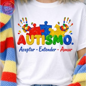 May include: White t-shirt with a colorful design featuring puzzle pieces and handprints. The word "AUTISMO" is displayed in vibrant colors, with the phrases "Aceptar, Entender, Amar" below. The shirt has a crew neck and short sleeves.