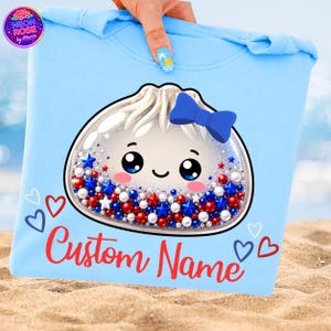 Könnte beinhalten: Hellblaues T-Shirt mit einem niedlichen Knödel-Design, einer blauen Schleife und roten, weißen und blauen Sternen und Perlen. Der Text "Custom Name" ist rot geschrieben. Das Shirt wird von einer Hand mit einem Nageldesign zum Thema Strand gehalten.