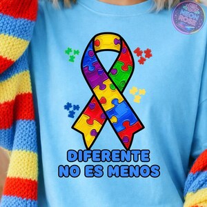 May include: Light blue sweatshirt featuring a colorful puzzle-piece ribbon design, with the text "DIFERENTE NO ES MENOS" in bold letters. The design includes green, yellow, red, purple, and blue puzzle pieces. A "Neon Rose" logo is visible in the upper right corner.