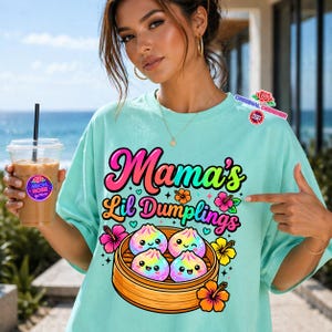 May include: A light teal t-shirt with the text "Mama's Lil Dumplings" in a colorful, playful font. The design features four smiling dumplings in a bamboo steamer, surrounded by flowers. A clear plastic cup with a straw is held in the left hand.