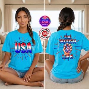 Könnte beinhalten: Ein hellblaues T-Shirt mit einem Grafikdesign auf der Vorder- und Rückseite. Die Vorderseite zeigt den Text USA in einer rot-weiß-blauen Farbverlaufsschrift mit Sternen und Feuerwerksakzenten. Die Rückseite zeigt eine Hand, die ein Spielzeug drückt, mit dem Text THE SQUISH LIFE.