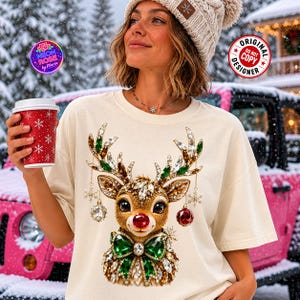 Könnte beinhalten: Ein cremefarbenes Kurzarm-T-Shirt mit einer Grafik eines Rentiers, das mit grünen und klaren Kristallen, einer roten Nase und einer grünen Kristallfliege verziert ist. Das Rentiergeweih ist mit hängenden Kristall-Schneeflocken und Kugeln geschmückt.