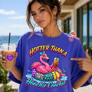 May include: A blue t-shirt with a graphic of a pink flamingo wearing a bow and holding a drink, floating on a colorful inner tube. The text reads "HOTTER THAN A Hoochie Coochie". A clear plastic cup with a drink is held in the left hand.