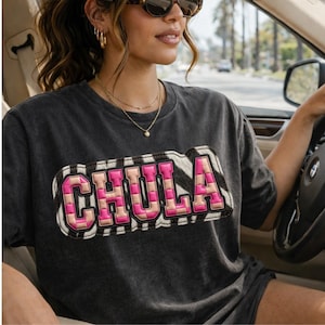 Può includere: T-shirt grigio scuro con la scritta "CHULA" in lettere rosa e beige a scacchi, bordata di bianco e stampa zebrata. La maglietta è girocollo e sembra essere realizzata in un materiale morbido.