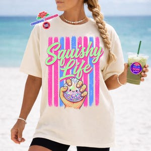 May include: Cream-colored t-shirt with the words "Squishy Life" in a neon green script, set against pink and blue vertical stripes. The shirt features a cartoon hand holding a squishy toy with a smiling face. The image includes the text "Original Designer" and "Do Not Copy".