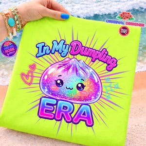 Puede incluir: Un cojín verde neón con el texto "In My Dumpling ERA" y una ilustración de dumpling kawaii. El dumpling es colorido con detalles de purpurina. El cojín lo sostiene una persona con esmalte de uñas azul y pulseras.