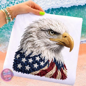 Puede incluir: Tela blanca con un bordado detallado de una cabeza de águila calva. La cabeza del águila tiene plumas blancas, un pico amarillo y una mirada severa. El diseño incorpora la bandera estadounidense.