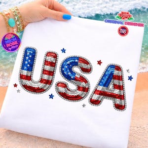 May include: White t-shirt with the word "USA" spelled out in red, white, and blue sequins. The letters are outlined in red sequins and surrounded by small star-shaped sequins. The shirt is held up by a hand.