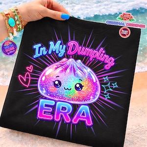 May include: Black t-shirt featuring a colourful graphic of a dumpling with kawaii eyes and the text "In My Dumpling Era". The design includes pink and blue neon accents, hearts, and star shapes. The shirt is a fun, playful design.
