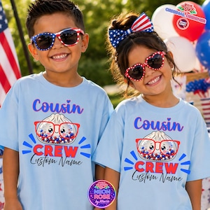 Könnte beinhalten: Zwei Kinder tragen hellblaue T-Shirts mit dem Wort "Cousin" in blauer Schrift. Die Shirts zeigen eine Grafik einer Teigtasche mit Brille und roten, weißen und blauen Akzenten. Der Text "CREW Custom Name" steht darunter.