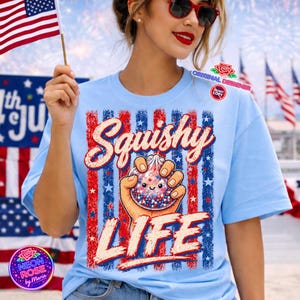 Könnte beinhalten: Hellblaues T-Shirt mit patriotischem Design. Das Shirt zeigt die Worte "Squishy LIFE" in Rot und Weiß, mit einer Grafik einer Hand, die ein Squishy-Spielzeug mit roten, weißen und blauen Sternen hält. Das Design umfasst rote und blaue Streifen und Sterne.