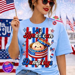 Könnte beinhalten: Hellblaues T-Shirt mit dem Text "All American Dumplings" in Rot und Blau. Das Design zeigt eine Teigtasche in einem Dampfkorb, eine Rakete, Sterne und ein amerikanisches Flaggenthema.
