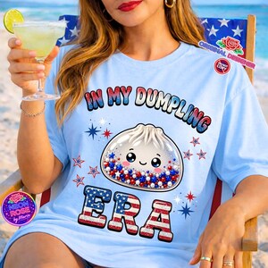 Könnte beinhalten: Hellblaues T-Shirt mit dem Text "IN MY DUMPLING ERA" und einer Abbildung eines Knödels mit roten, weißen und blauen Sternen. Das Shirt hat eine lockere Passform und ist für Freizeitkleidung geeignet.