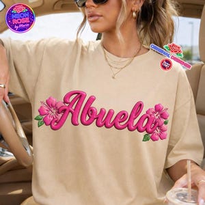 May include: A beige t-shirt featuring the word "Abuela" in vibrant pink, adorned with hibiscus flowers. The design includes the text "Neon Rose by Maria" and "Original Designer" in a circular logo.