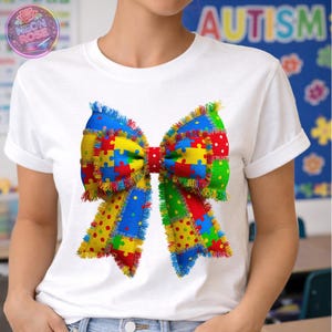 May include: White t-shirt featuring a large, colorful bow design. The bow is made of puzzle pieces in red, yellow, blue, and green, with a red polka-dot center and fringed edges. The word "AUTISM" is visible in the background.