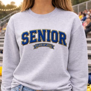Könnte beinhalten: Hellgraues Sweatshirt mit dem Wort "SENIOR" in blauen und goldenen Buchstaben. Darunter steht der Text "CLASS OF 2026" in einem Banner. Das Sweatshirt hat einen Rundhalsausschnitt.