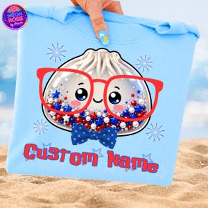 Könnte beinhalten: Hellblaues T-Shirt mit einer Cartoon-Knödel-Figur, die eine rote Brille und eine blaue Fliege trägt. Der Knödel hat ein lächelndes Gesicht und ist mit roten, weißen und blauen Sternen und Perlen gefüllt. Der Text "Custom Name" ist rot gedruckt.