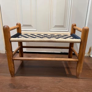 May include: A small wooden bench with four rounded legs and a woven seat featuring a geometric pattern in cream and dark blue. The frame is constructed from light brown wood with horizontal support bars connecting the legs on each side.