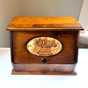 May include: A wooden bread box with a dark brown finish. The front features an oval carving of kitchen utensils. A drawer is located at the bottom with a round wooden knob. The box is designed to store bread and baked goods.