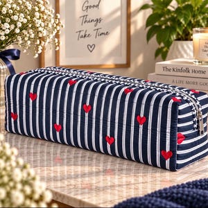 May include: A rectangular quilted pencil case featuring a navy blue and white vertical striped pattern with small embroidered red hearts scattered across the surface. The case includes a silver metal zipper closure along the top edge.