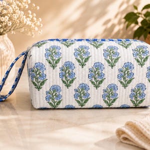 May include: A rectangular quilted cosmetic bag featuring a white base with a repeating pattern of blue flowers and green stems. The bag includes a blue and white striped fabric wrist strap attached to the side and a matching striped zipper trim.
