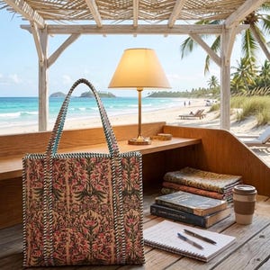 May include: A patterned tote bag with a floral design and green trim stands on a wooden desk. A brass lamp, books, a notepad, and a coffee cup are also present. The background shows a beach with palm trees and turquoise water.